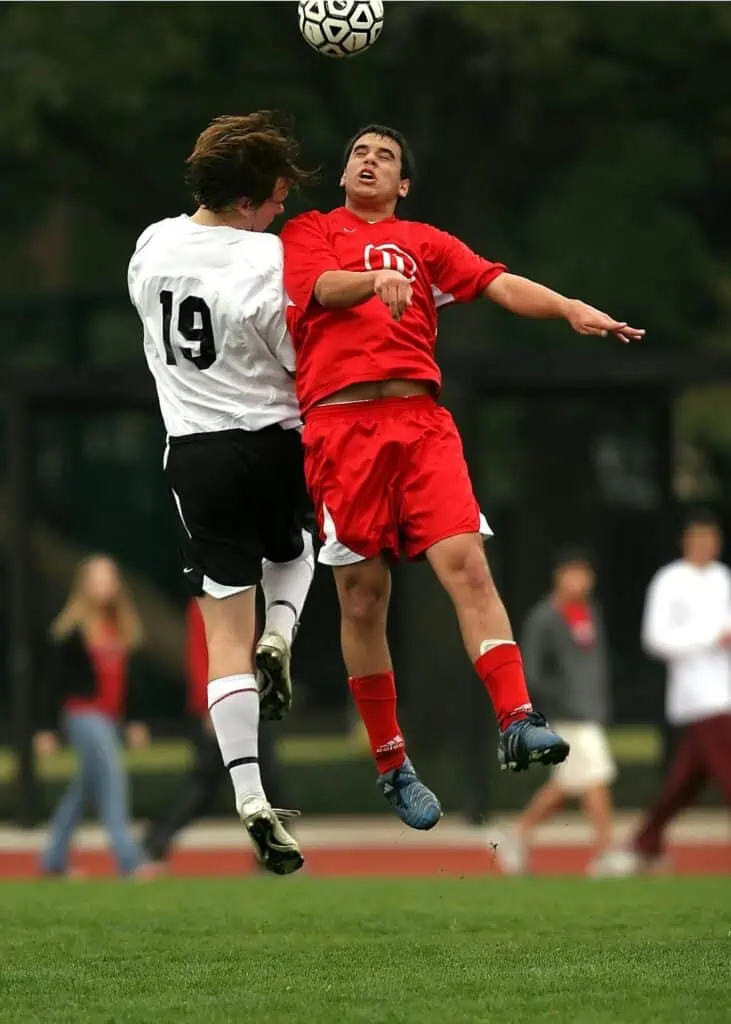 To fodboldspillere, den ene i en hvid trøje og den anden i en rød trøje, hopper i luften og konkurrerer om en bold under en kamp. alt-om-fodbold.dk